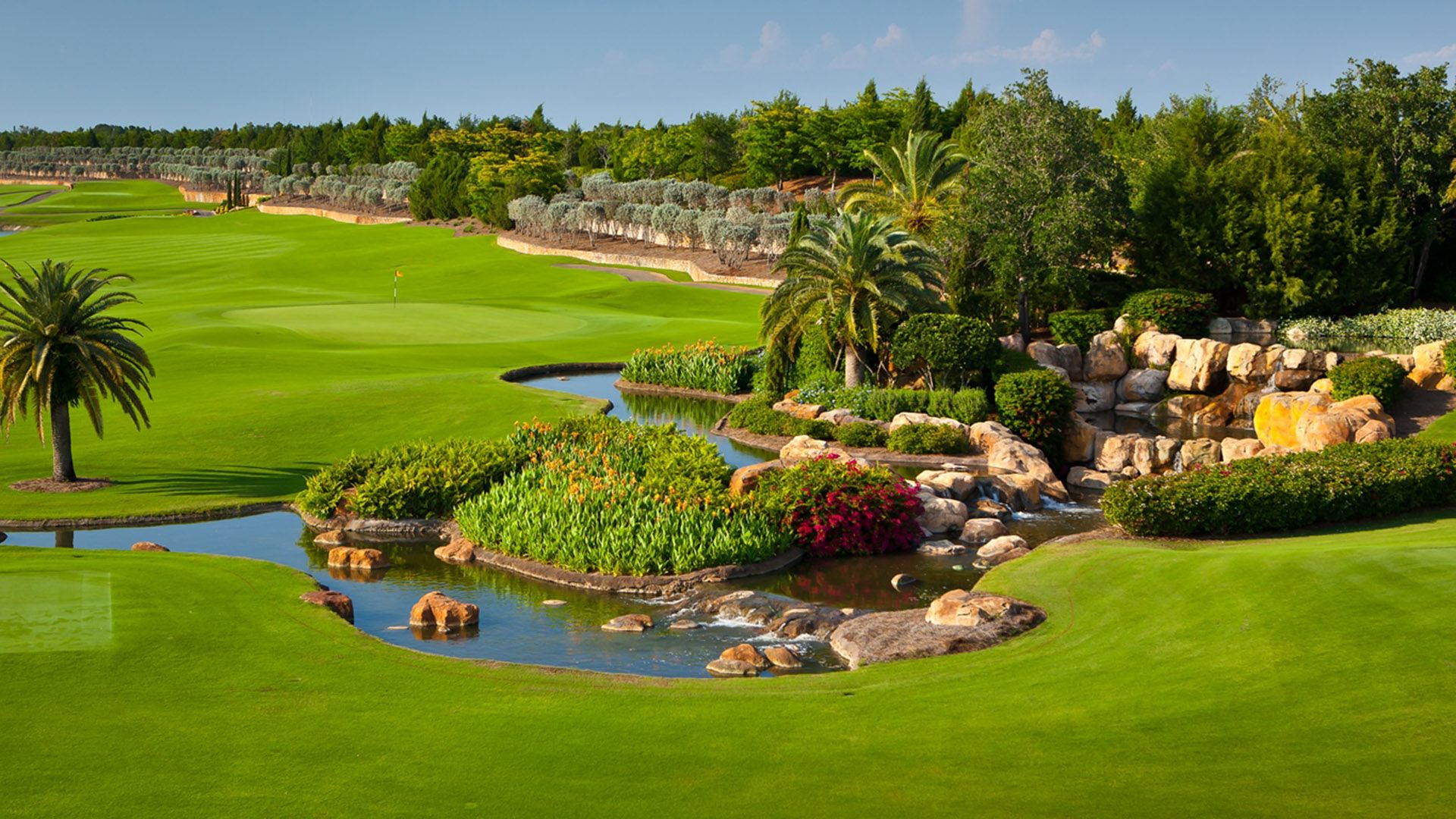 Lush golf course with a manicured green, surrounded by palm trees, vibrant flowerbeds, a small stream, and cascading rocks under a clear sky.