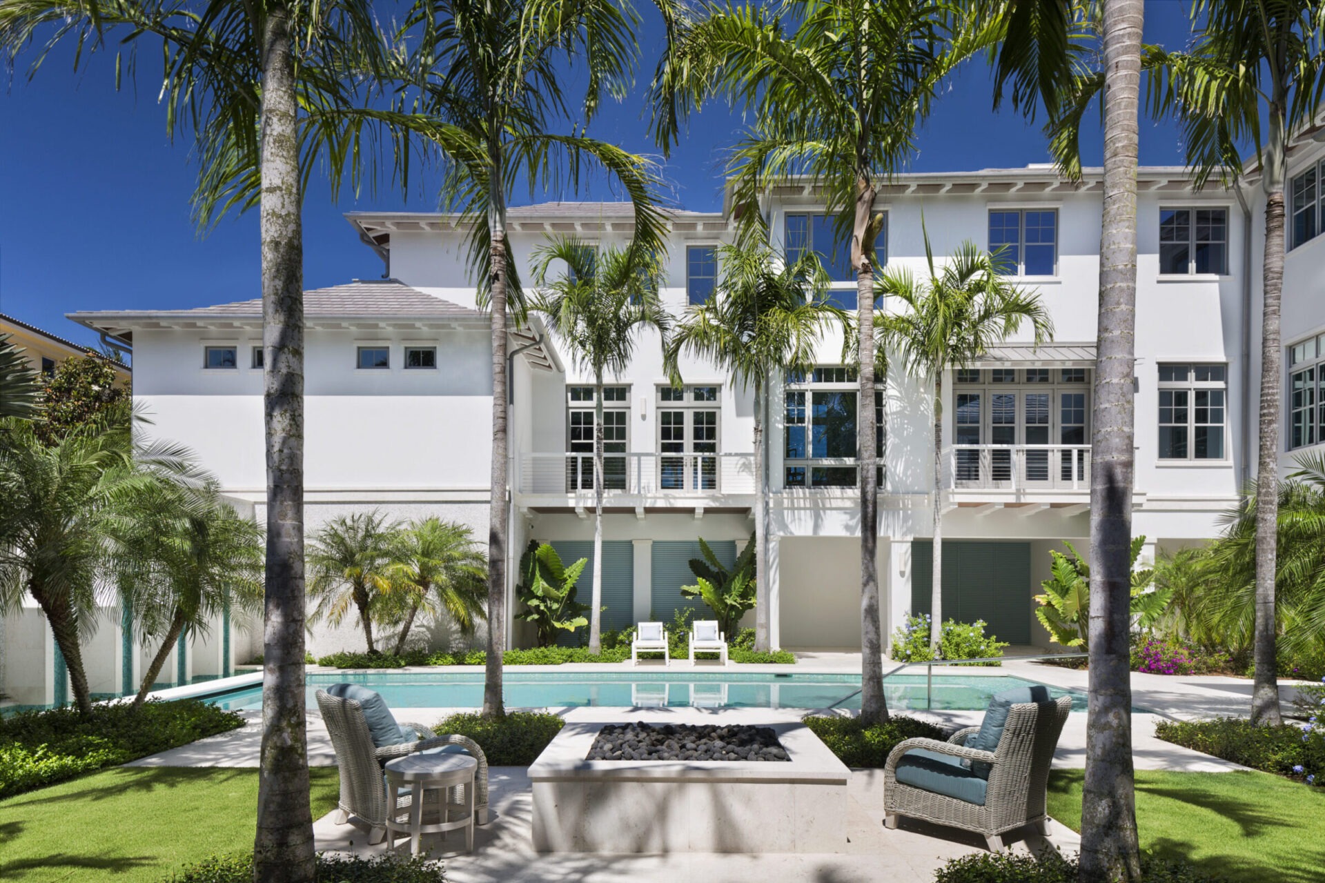 A modern white house with palm trees, pool, and patio furniture under a bright blue sky. Luxurious outdoor setting; serene ambiance.