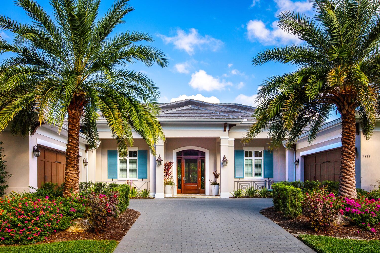 A modern house with blue shutters, palm trees, and vibrant flowers under a clear blue sky. Paved driveway leads to the entrance.