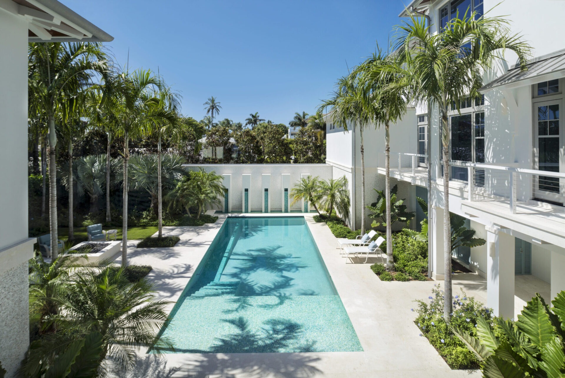 Luxurious pool surrounded by tropical plants, modern white building, and deck chairs. Clear blue sky enhances the serene, idyllic atmosphere.