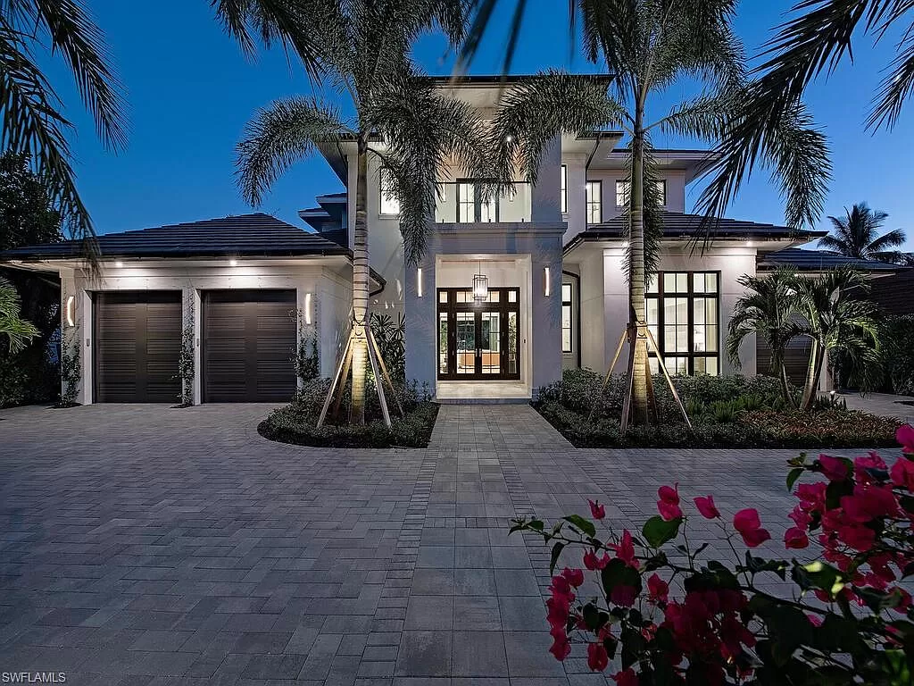 Modern two-story house with palm trees, well-lit at dusk. Spacious driveway and garage. Vibrant pink flowers in the foreground enhance elegance.