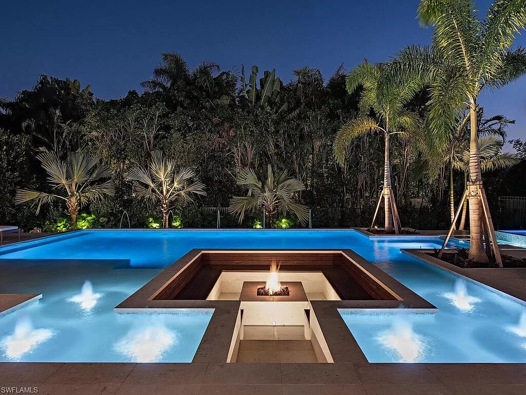 Luxurious pool with central fire pit surrounded by palm trees, lit beautifully against a night sky. Landscaped foliage frames the serene setting.