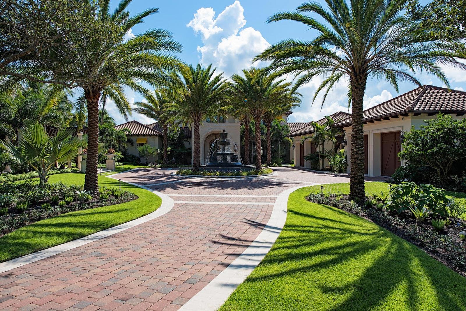 A luxurious villa entrance features palm trees, manicured lawns, and a central fountain, creating an inviting, tropical ambiance under a bright sky.