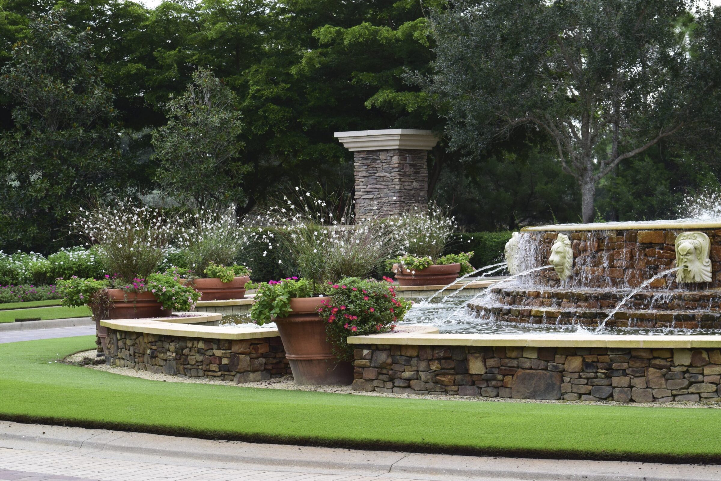 A landscaped garden with a stone fountain, featuring lion head spouts and surrounded by potted plants and lush greenery.