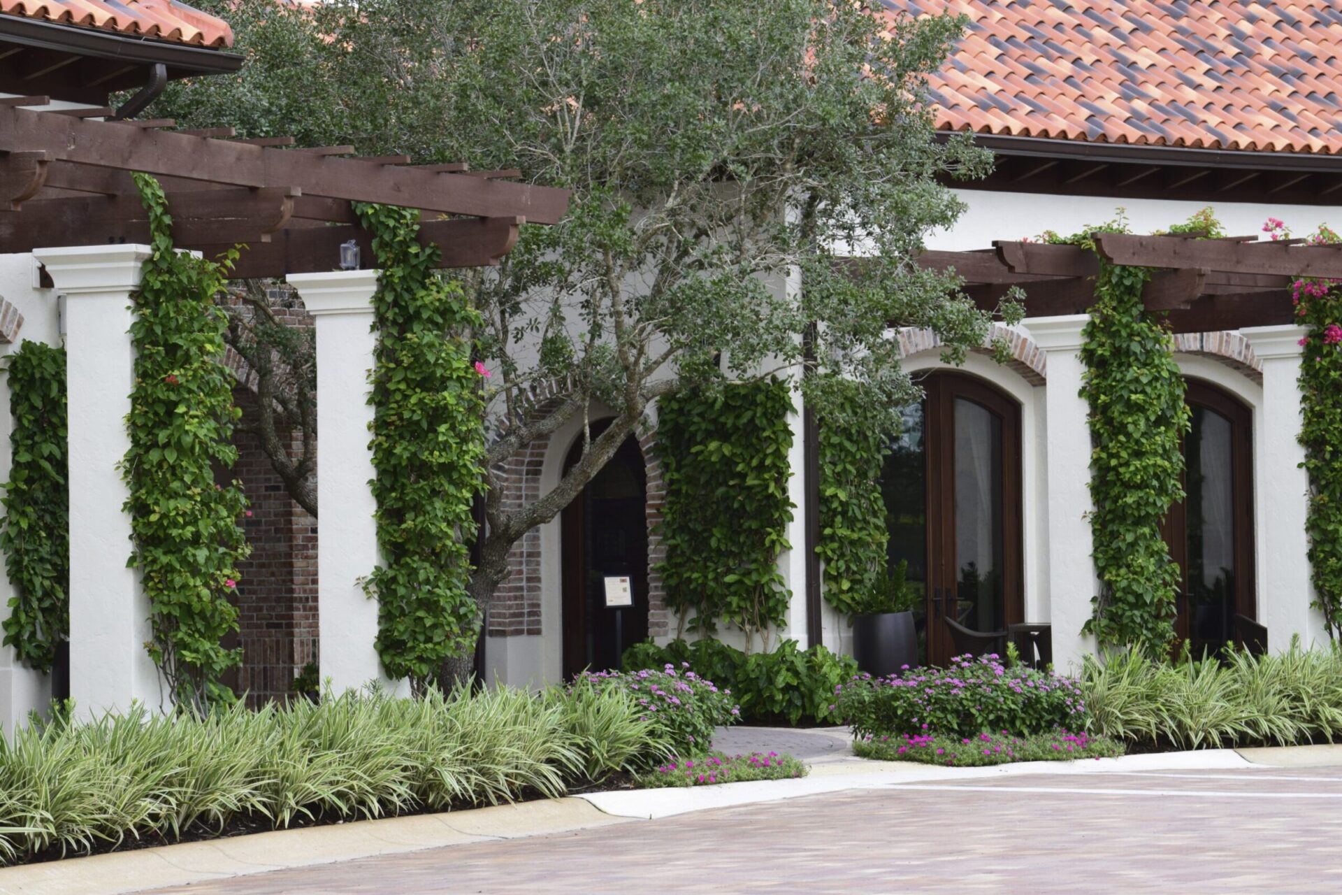 The image shows a building with terracotta roof tiles, lush greenery climbing white columns, arched windows, and well-maintained surrounding plants.