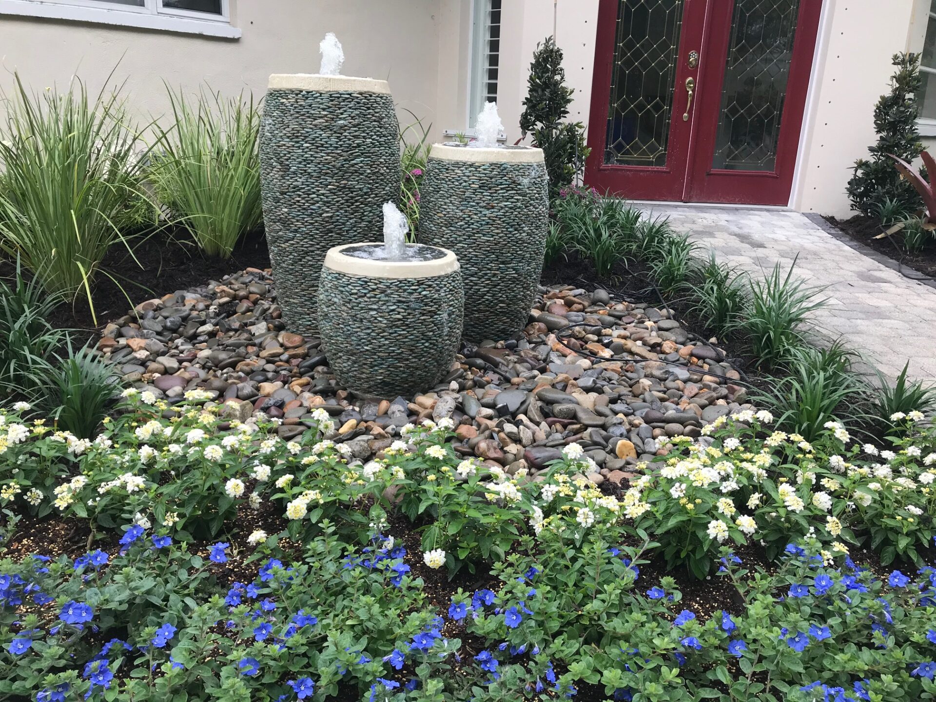A landscaped garden features three tiered water fountains surrounded by colorful flowers, pebbles, and greenery near a building with red double doors.