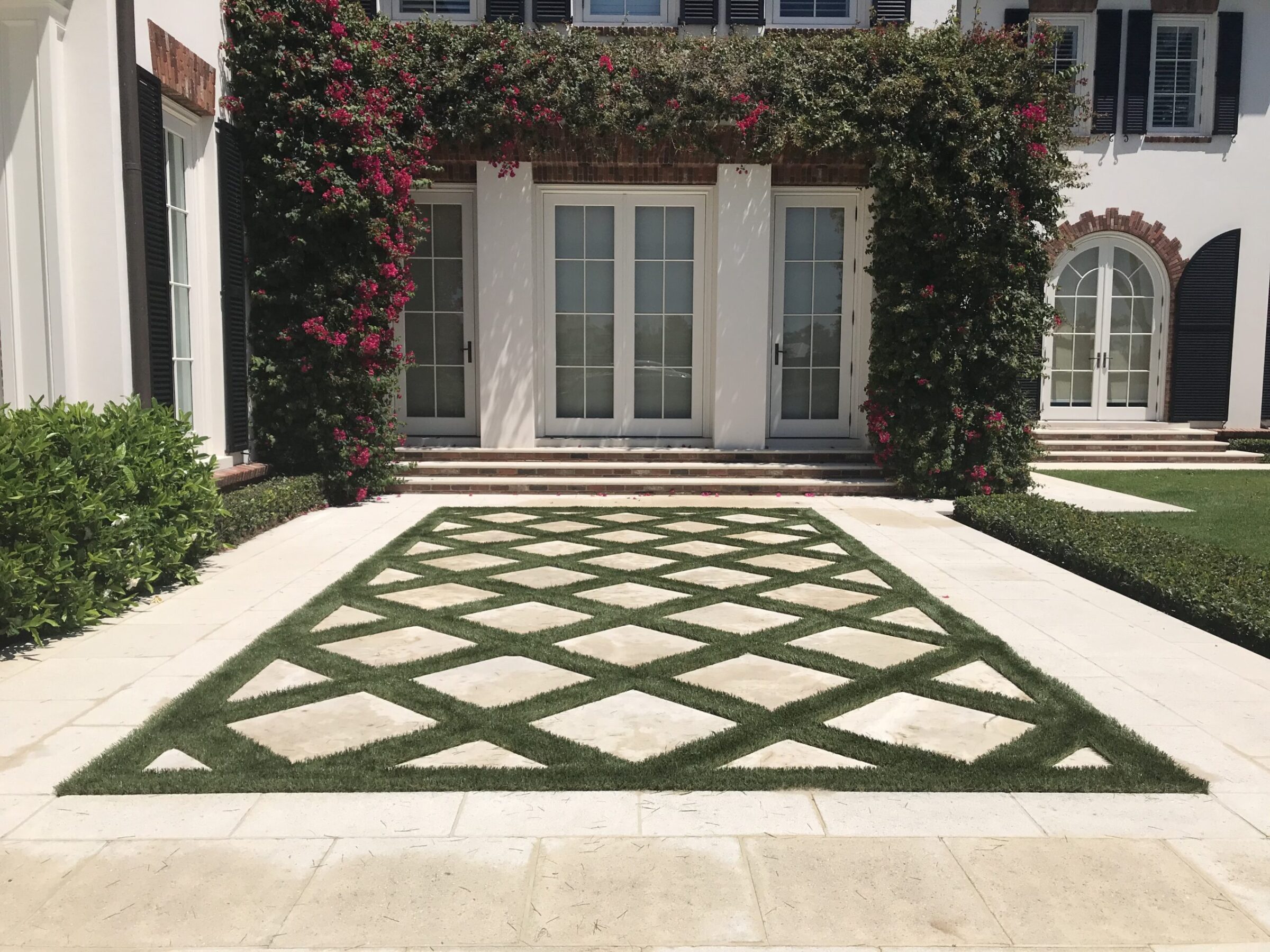 Elegant house facade with greenery and flowers climbing walls, framed by stunning geometric paving. Bright light highlights the lush, symmetrical landscaping.
