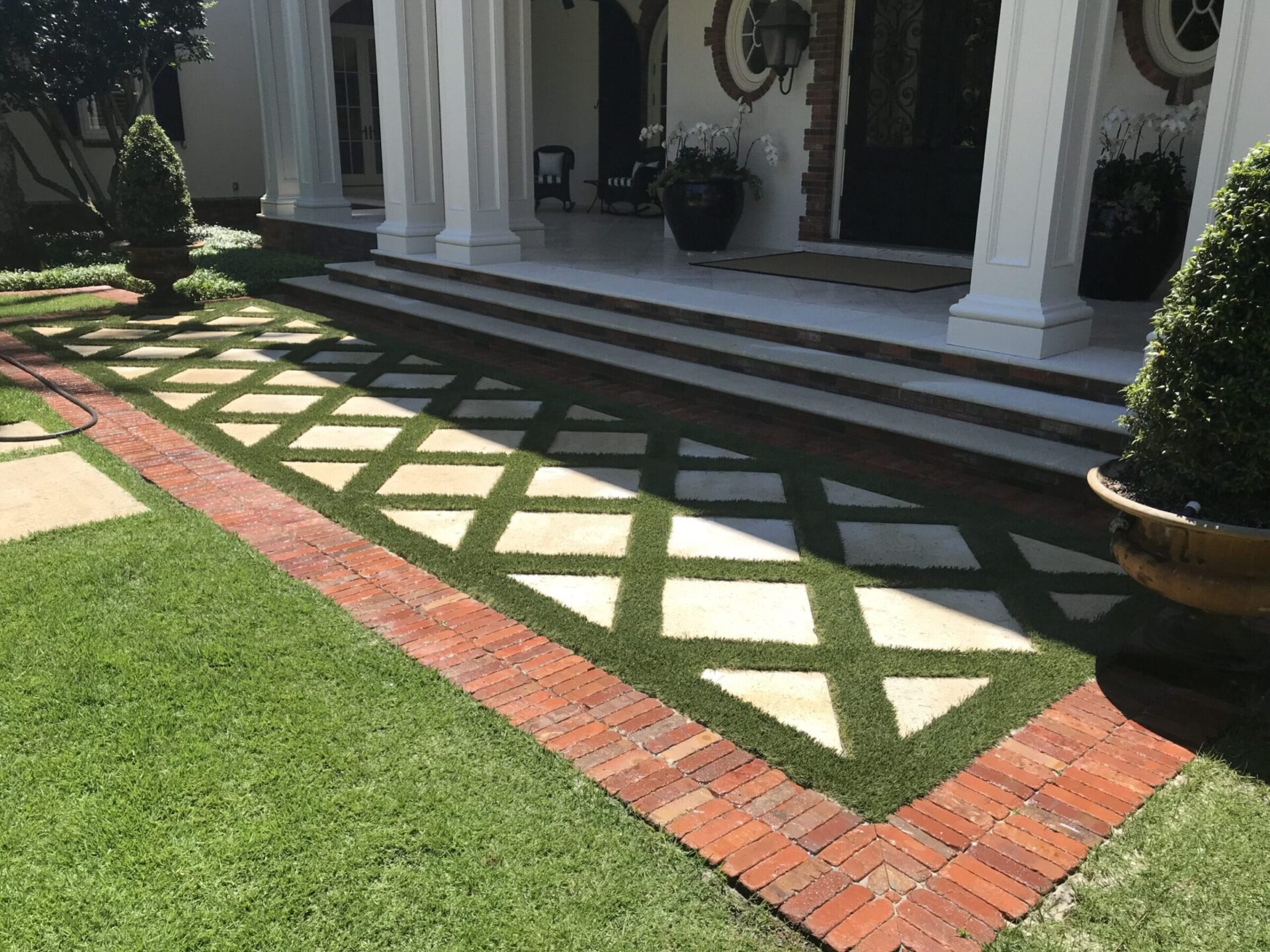 Grand entrance with geometric pathway, brick borders, and manicured garden. Stately columns frame the entrance, complementing ornate doors and lush greenery.
