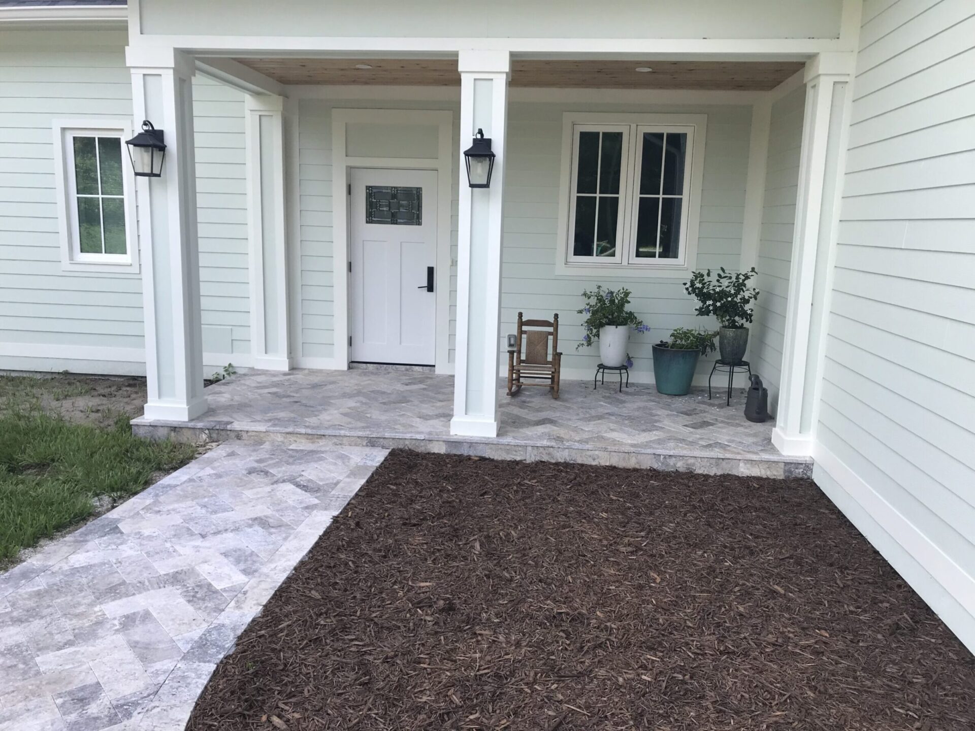 Front porch of a light-colored house with columns, potted plants, and a small chair. Mulch and a stone pathway lead to the entrance.