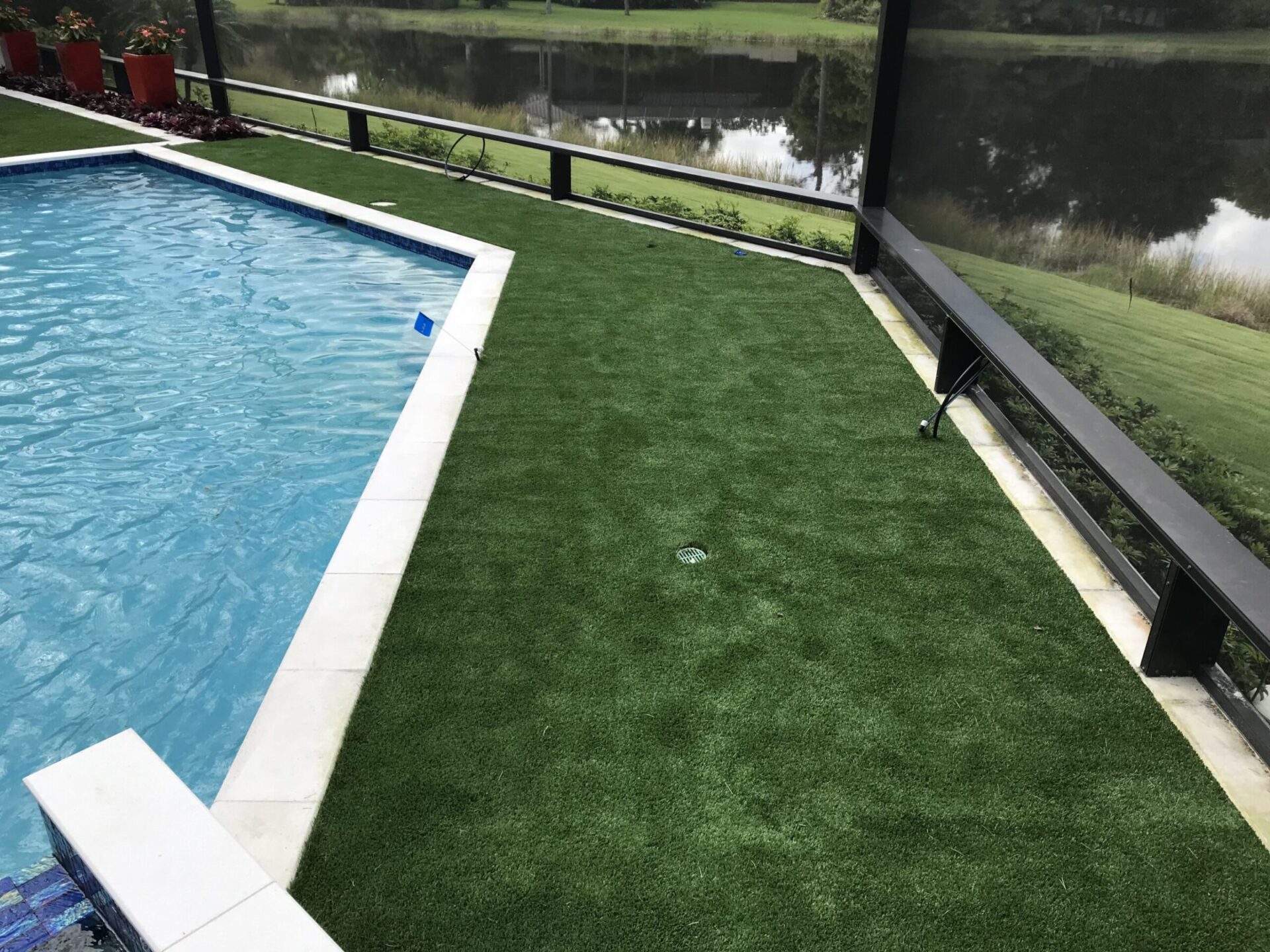 A pool with adjacent artificial grass overlooks a tranquil pond, bordered by potted plants and screened fencing, under a clear sky.