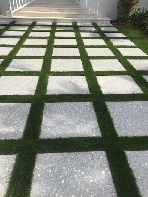 Geometric garden path of alternating gray tiles and green grass, leading up to white-railing, concrete stairs. No landmarks or historical buildings visible.
