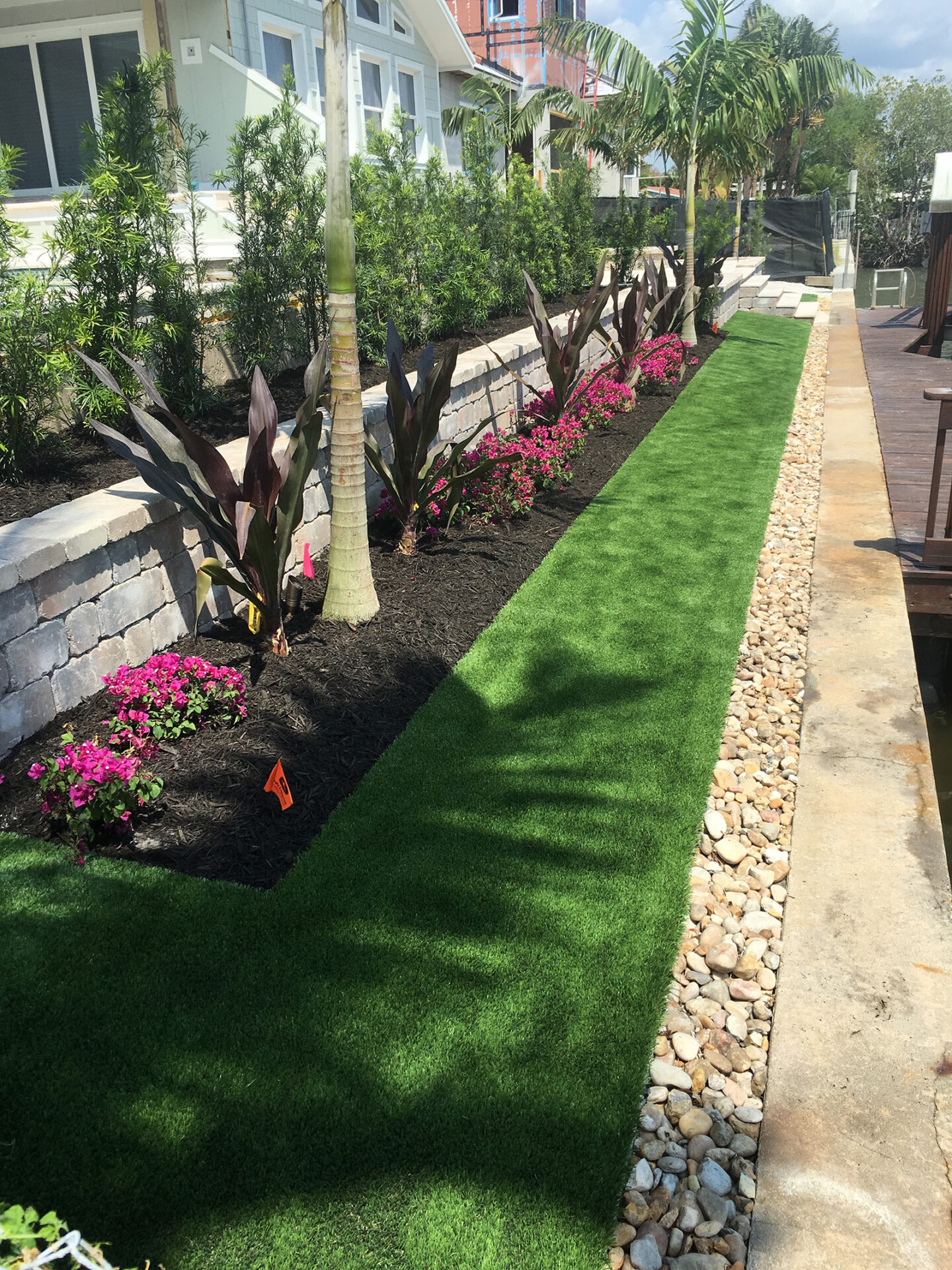 Side yard with vibrant green grass, tropical plants, and pink flowers next to a modern house. Stone path borders the landscaped area.