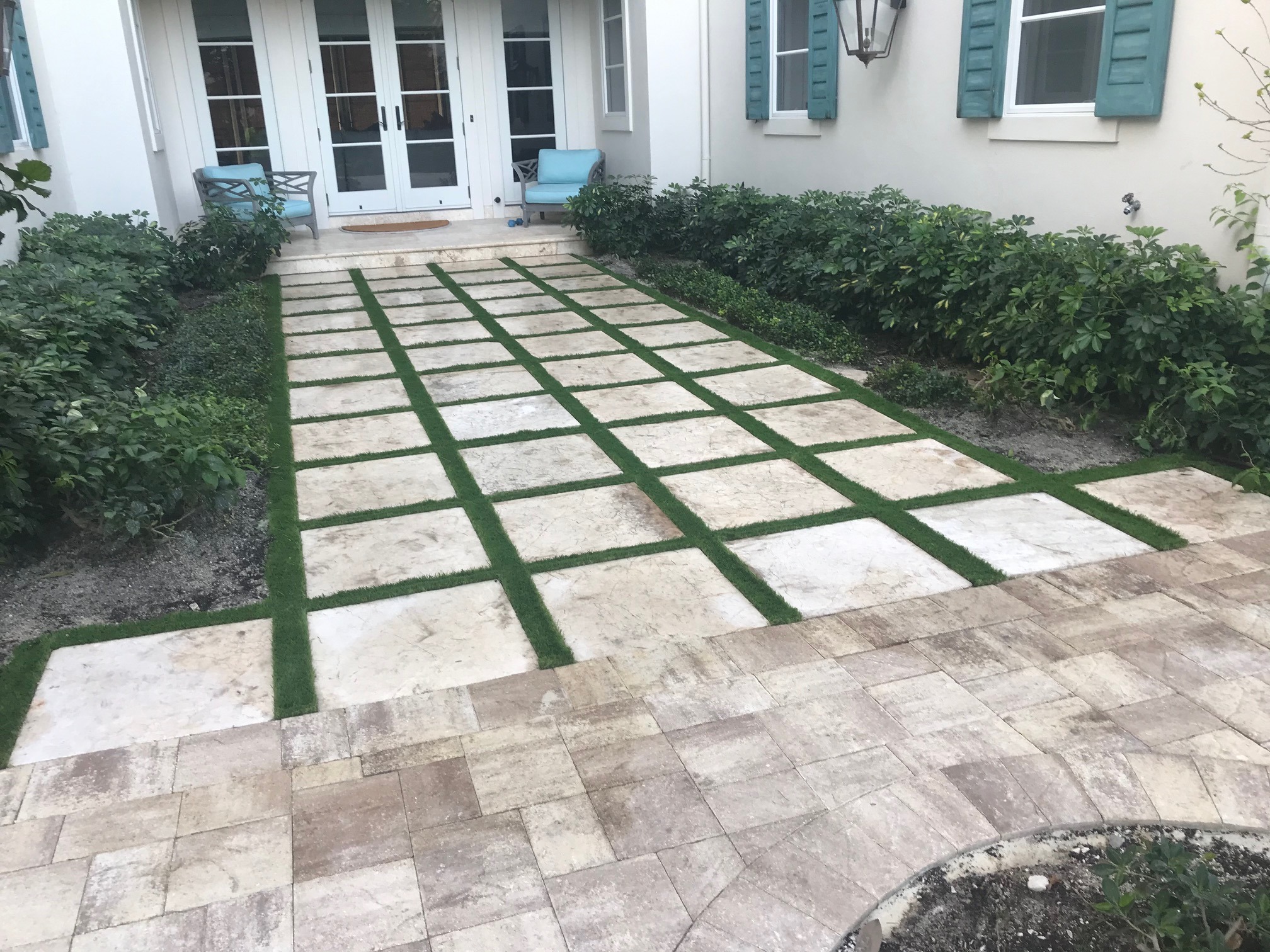 A courtyard features a travertine walkway with grass inlays, surrounded by shrubs. Blue chairs rest near a white door with glass panels.