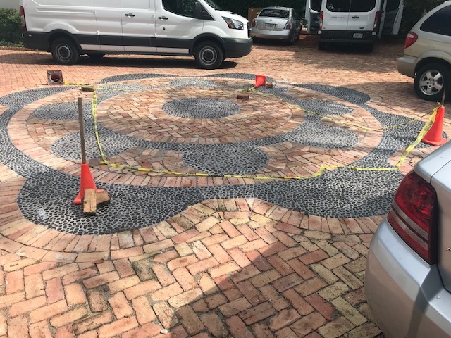 Brick and stone circular pattern on a driveway, surrounded by parked cars and traffic cones. No recognizable landmarks or historical buildings visible.