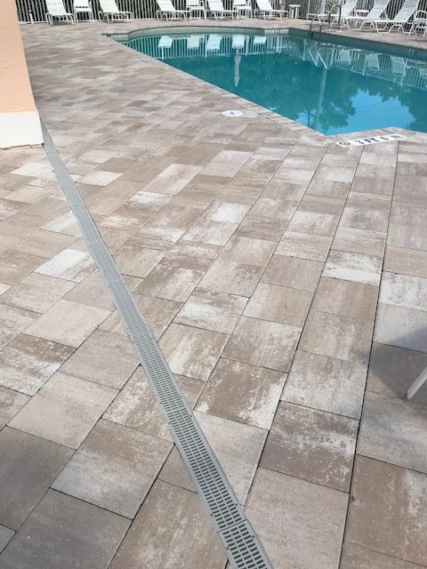 Outdoor pool area with beige tile flooring, surrounded by white lounge chairs. Calm blue water is reflected against the bright, sunny sky.