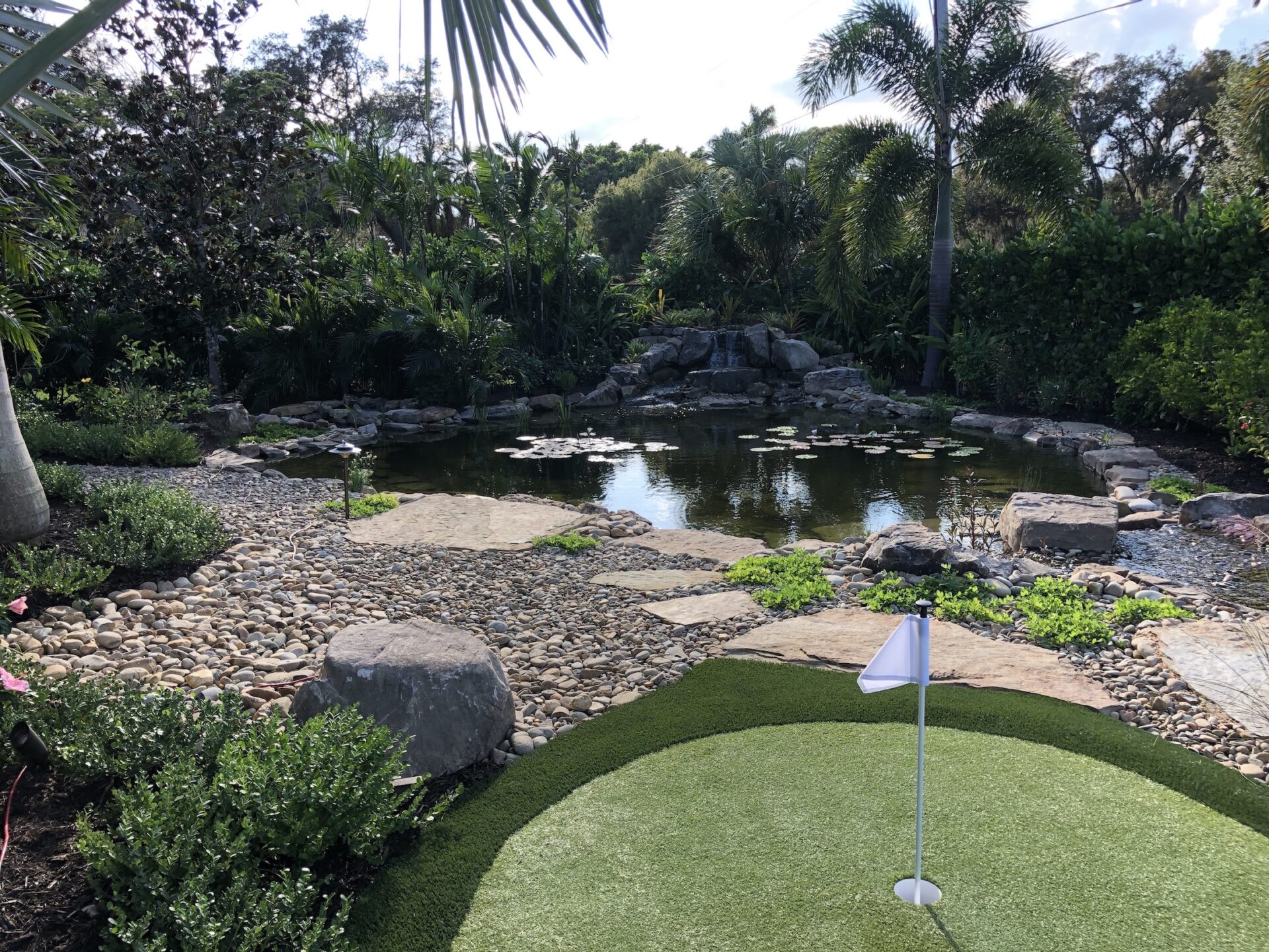 Lush garden scene features a rocky pond surrounded by palm trees, with a small putting green and flag in the foreground.