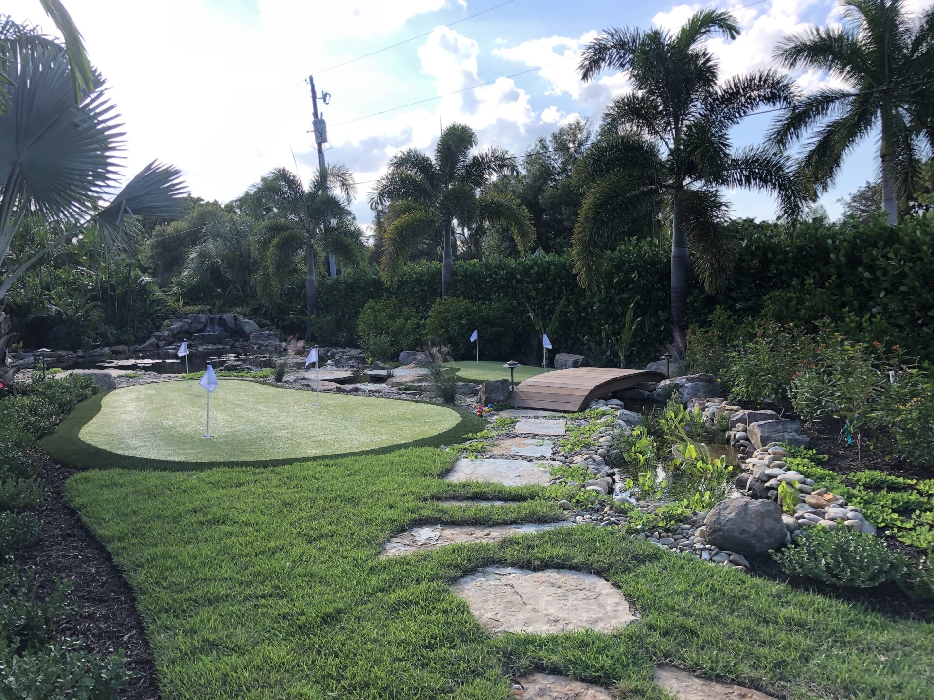 Lush garden scene with palm trees, a manicured putting green, small wooden bridge, and stone pathway surrounded by greenery and flowers.