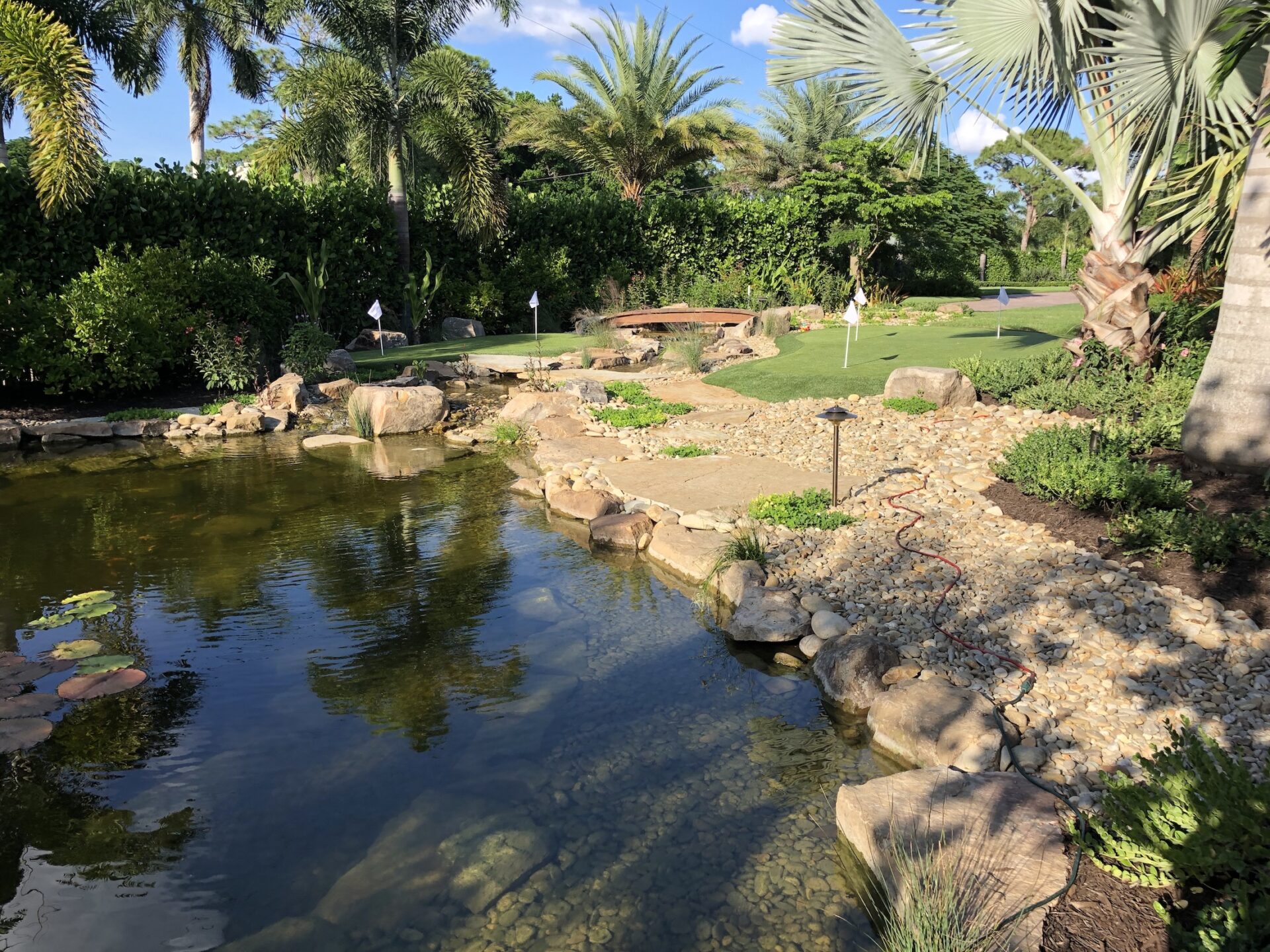 A serene garden scene features a pond, lush greenery, palm trees, and a small putting green with flagsticks under a clear blue sky.