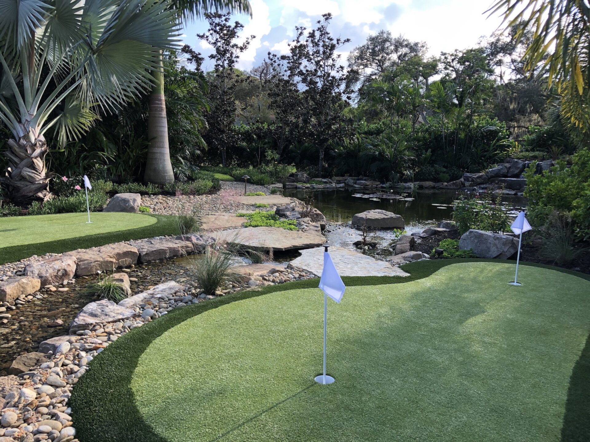 A serene mini-golf course with putting greens, white flags, lush greenery, and a nearby pond surrounded by rocks and palm trees.
