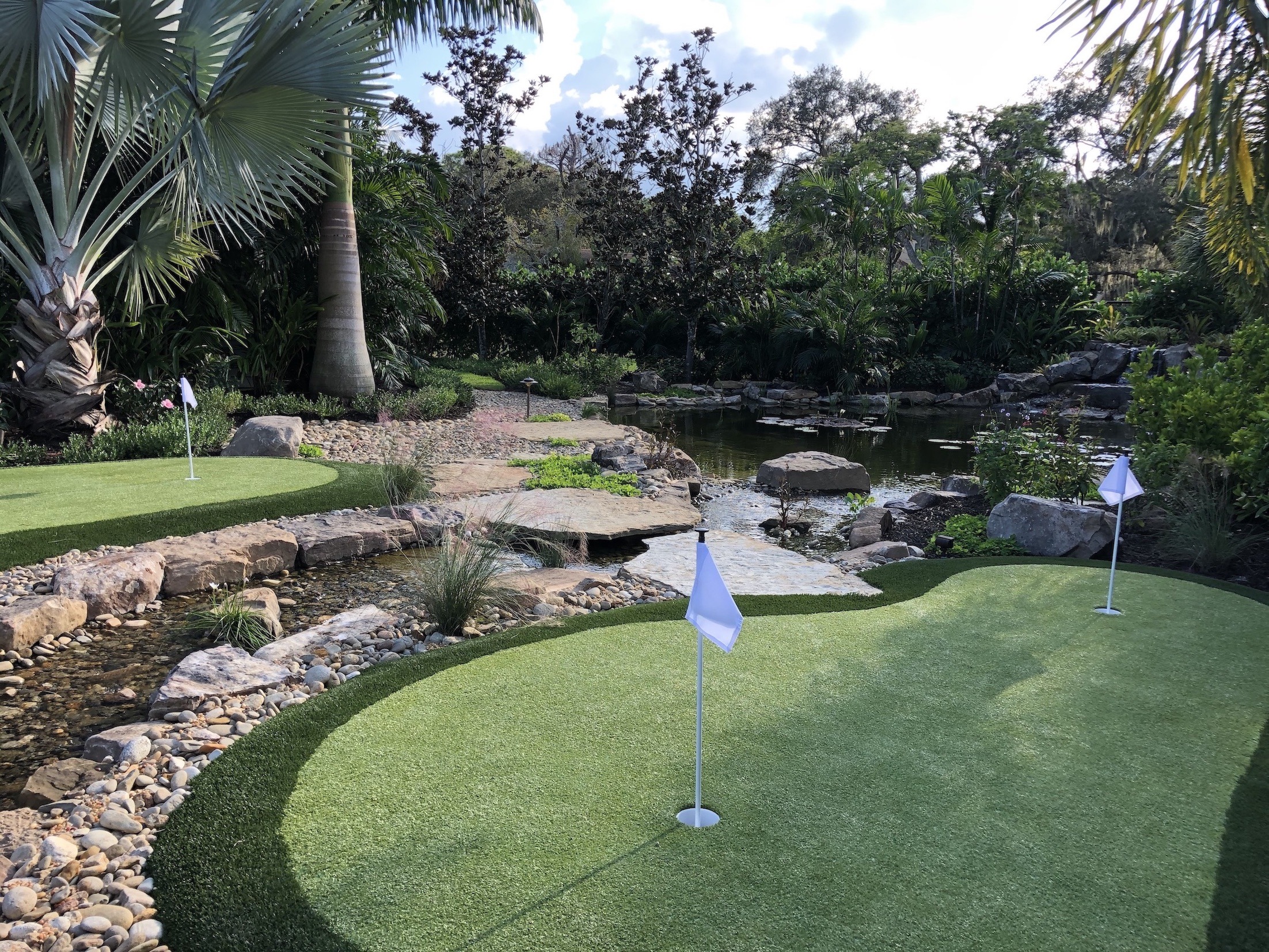 A serene mini-golf course with putting greens, white flags, lush greenery, and a nearby pond surrounded by rocks and palm trees.