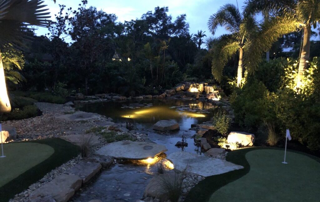 Illuminated garden scene at dusk with palm trees, a pond, rocks, and two small putting greens. Peaceful and serene setting.