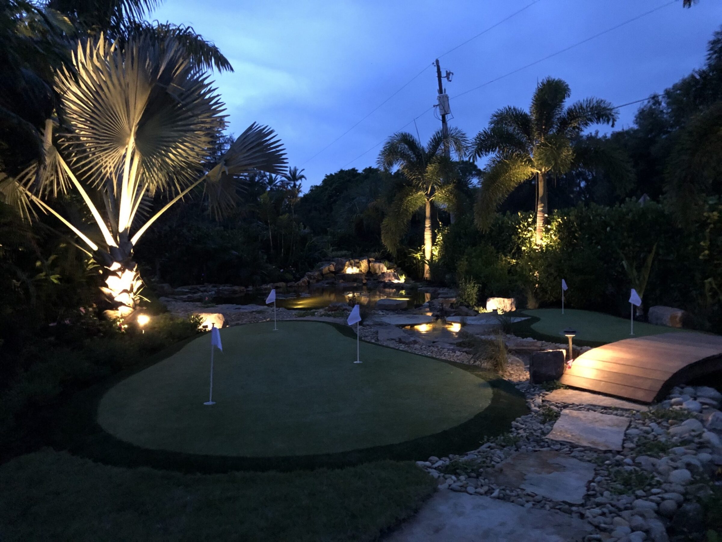 A serene night garden scene featuring a gently lit putting green, palm trees, a small bridge, and a tranquil water feature.
