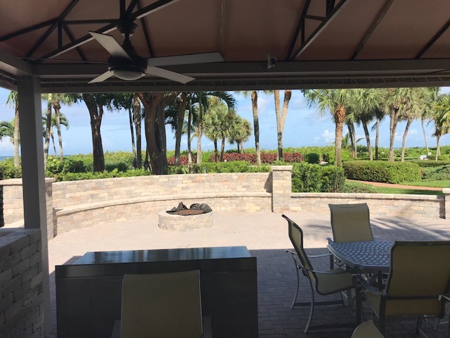 Covered patio with chairs and a table overlooks a garden with palm trees and a circular fire pit, under a bright blue sky.