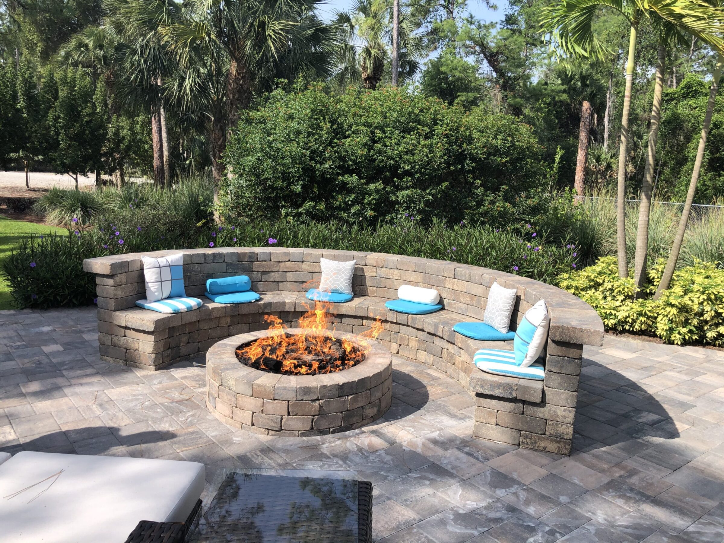 Curved stone seating around a blazing fire pit, with turquoise cushions. Surrounded by lush greenery, palm trees, and stone paving.