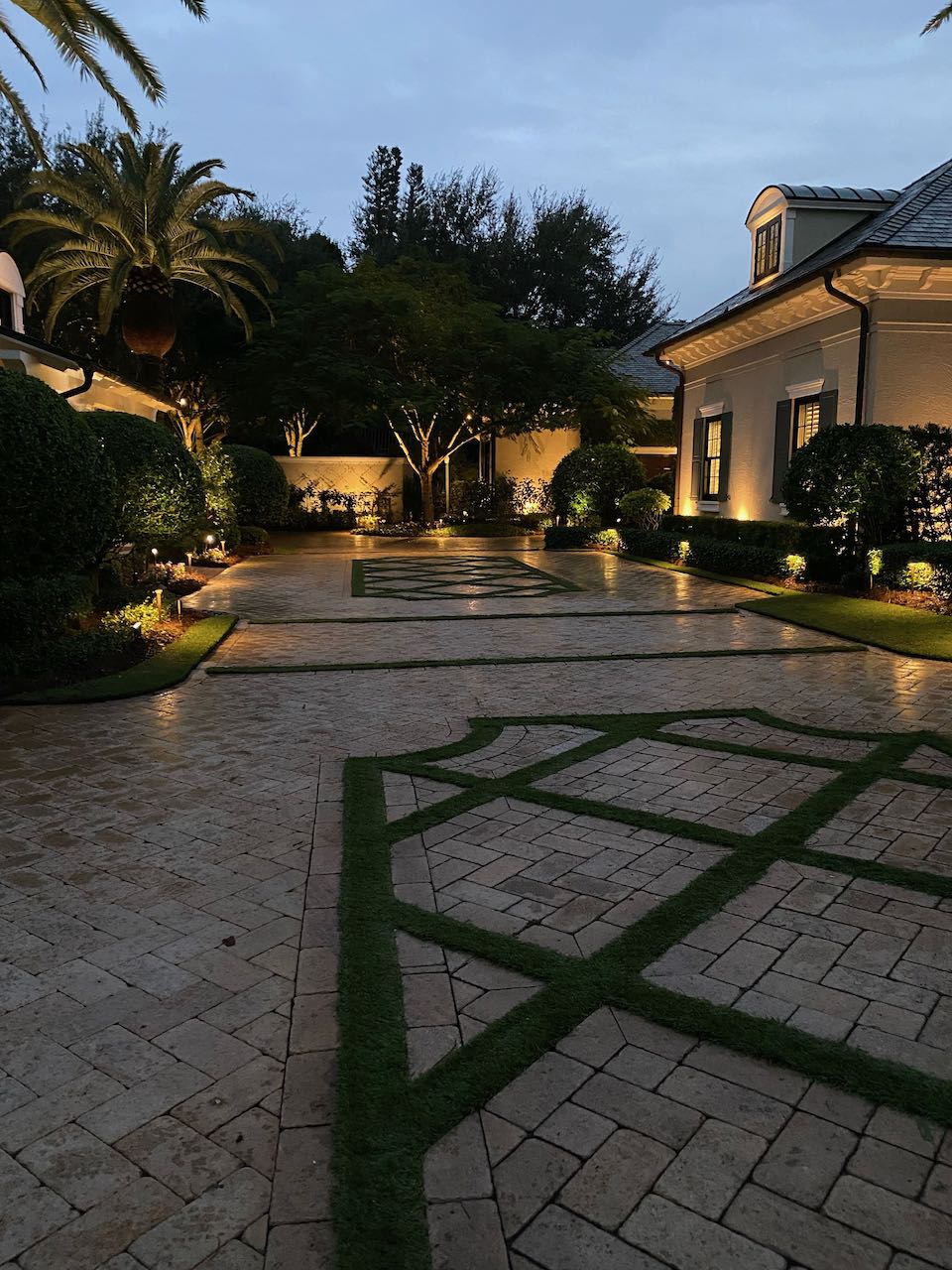 Elegant courtyard at twilight features manicured hedges, palm tree, geometric stone pathways, and subtle lighting, framed by traditional, upscale architecture.