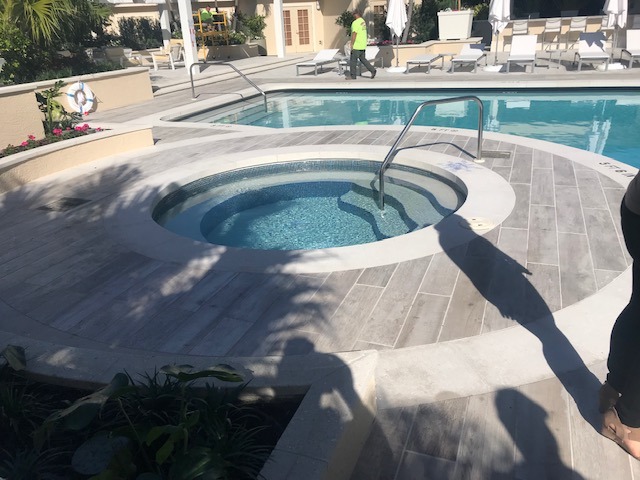 A sunlit pool area with a circular hot tub, lounge chairs, and a person in a green shirt walking nearby.