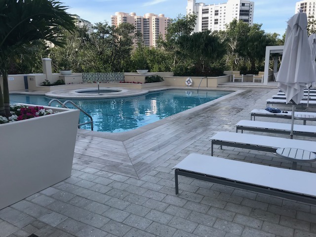 A serene swimming pool area with lounge chairs, surrounded by lush greenery and tall buildings in the background under a clear blue sky.