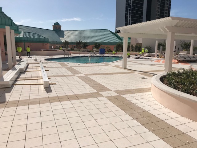 A swimming pool area under construction with several persons working. Green-topped buildings and a tall structure are visible in the background.