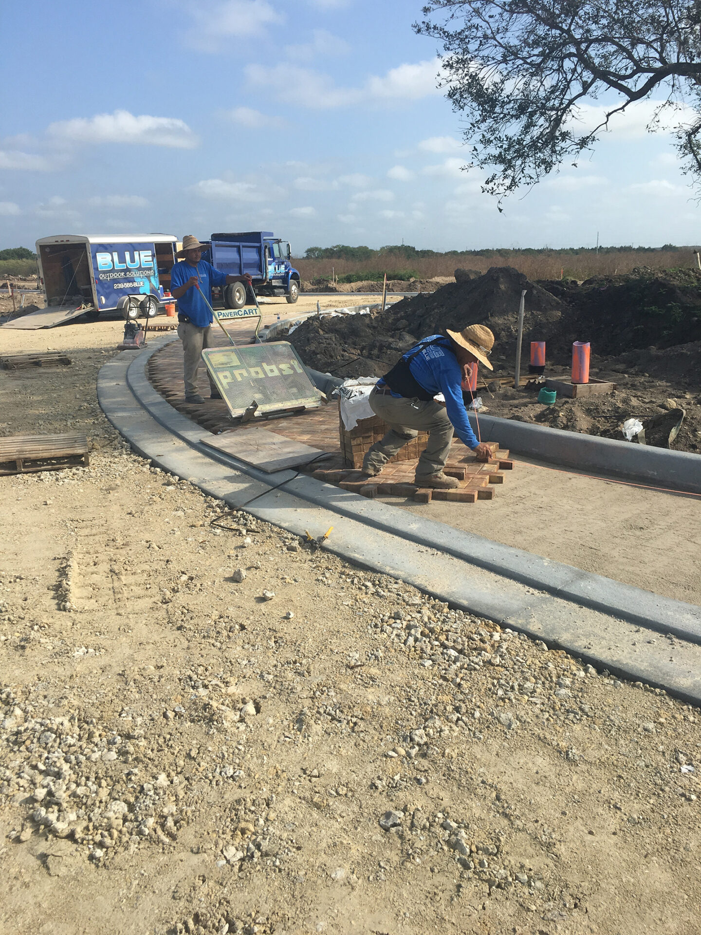 Two people lay bricks on a road construction site, with trucks and equipment visible under a clear sky. Tree in the background.