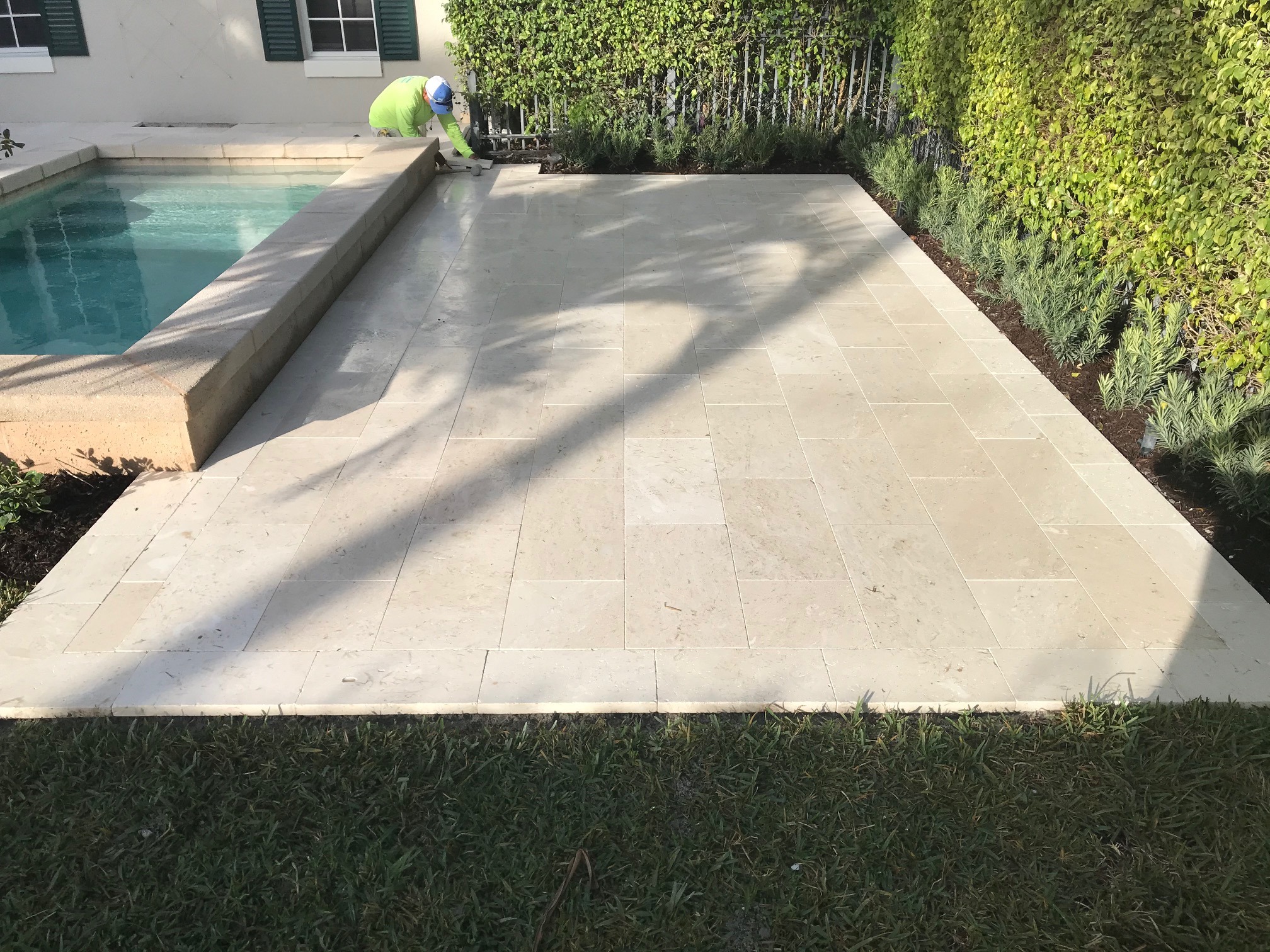 A person works on poolside tiling in a backyard, surrounded by lush greenery and a small swimming pool.