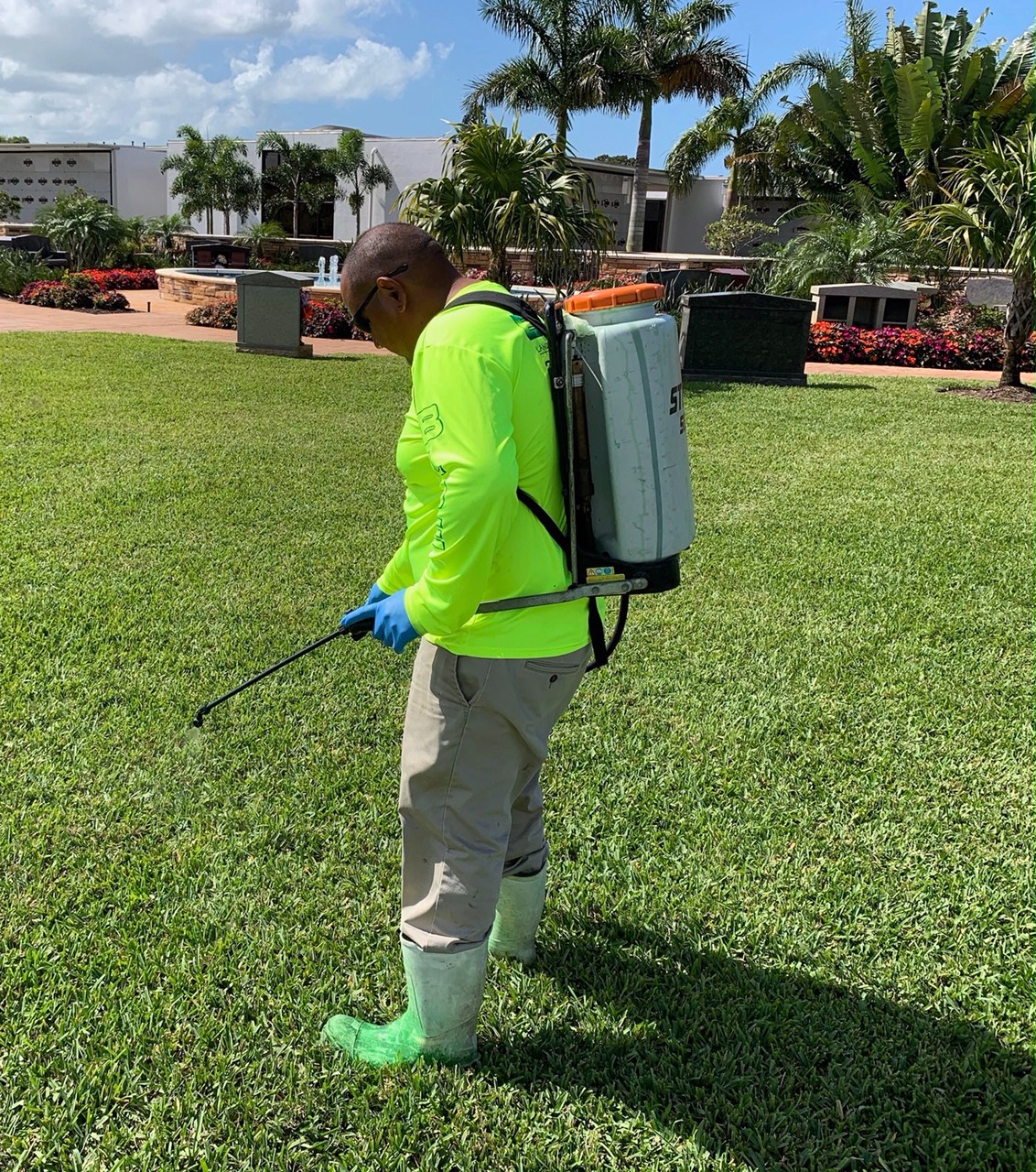 A person in bright clothing sprays a garden area with a backpack sprayer near palm trees and a landscaped building under a clear sky.