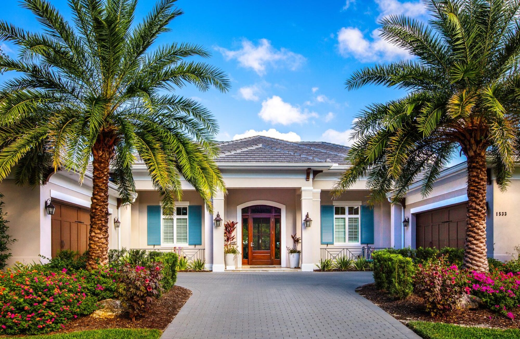 A modern, single-story house with palm trees and colorful flowers in front, under a clear blue sky, conveys a tropical suburban setting.