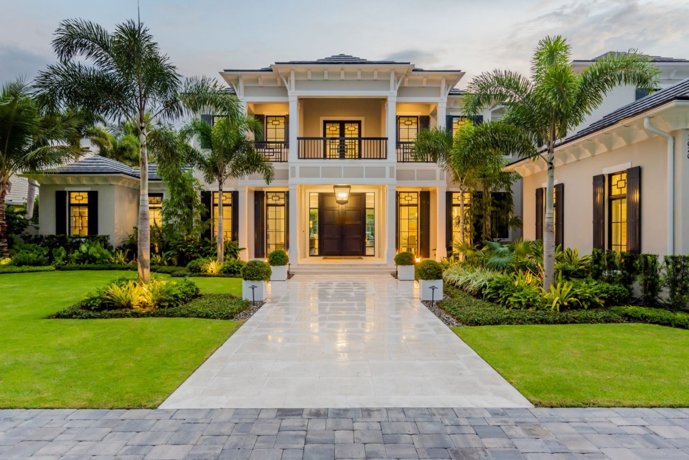 A large, elegant home with palm trees and manicured lawn. Symmetrical architecture and warm lighting enhance its grand entrance and inviting atmosphere.