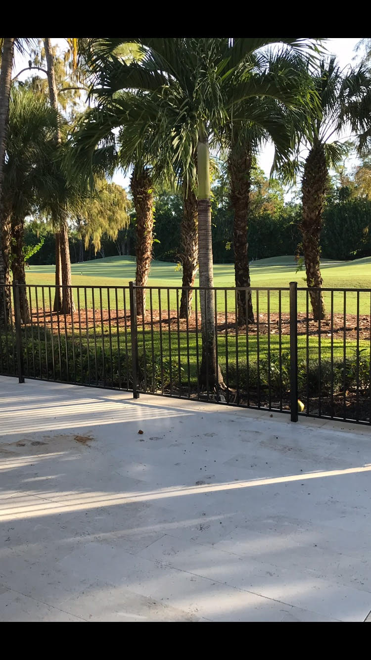 A sunny patio overlooks a lush golf course lined with palm trees, enclosed by a black metal fence. No landmarks are visible.