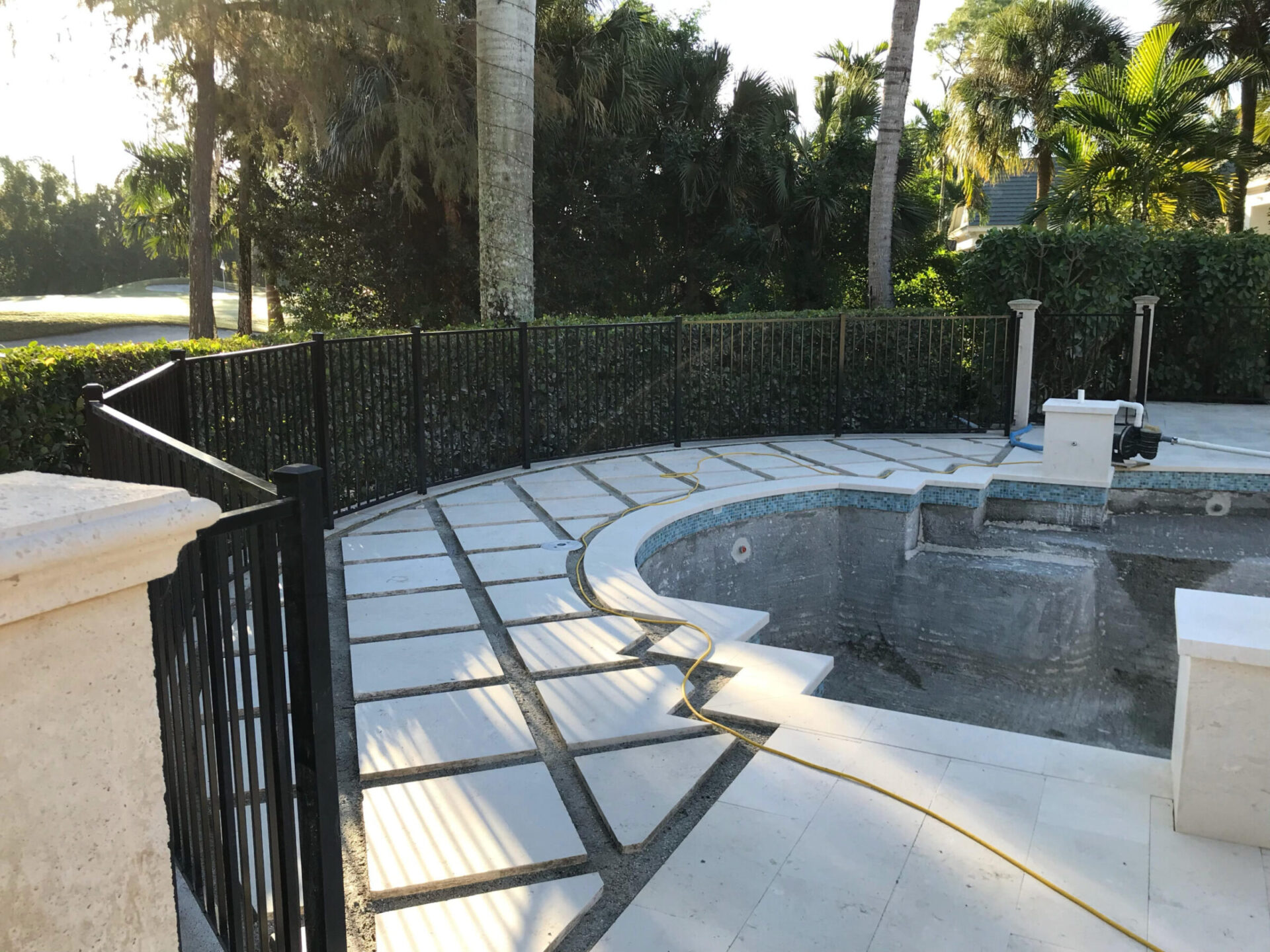 Empty swimming pool with surrounding black metal fence, tile paving, and lush greenery in the background. Yellow hose rests on the ground.
