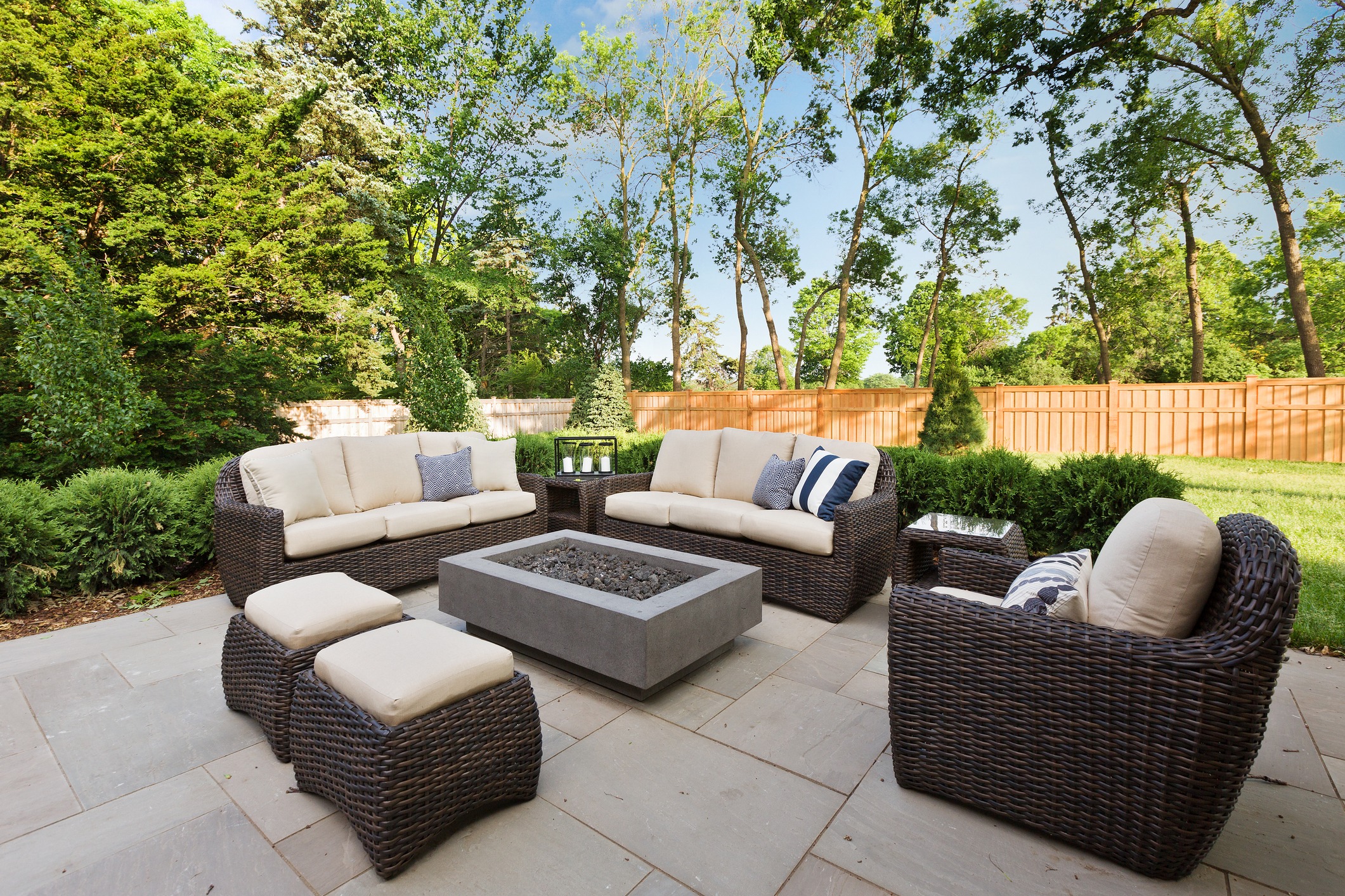 Cozy outdoor patio with wicker furniture, cushions, and a central fire pit. Surrounded by trees and a wooden fence, under a clear sky.