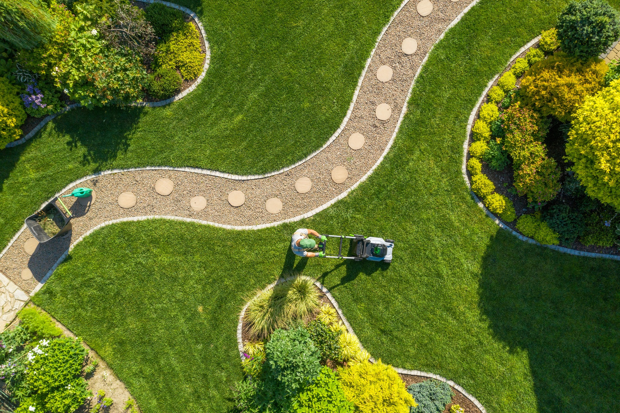 Aerial view of a landscaped garden with a person mowing the lawn, featuring curved pathways and vibrant green foliage.