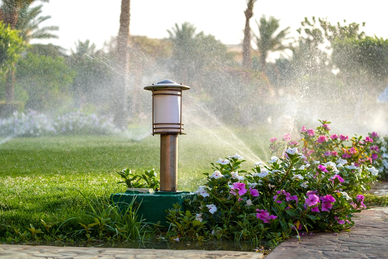 A garden with flowers, sprinklers, and a lamp post, surrounded by lush greenery and palm trees in a vibrant, sunny setting.