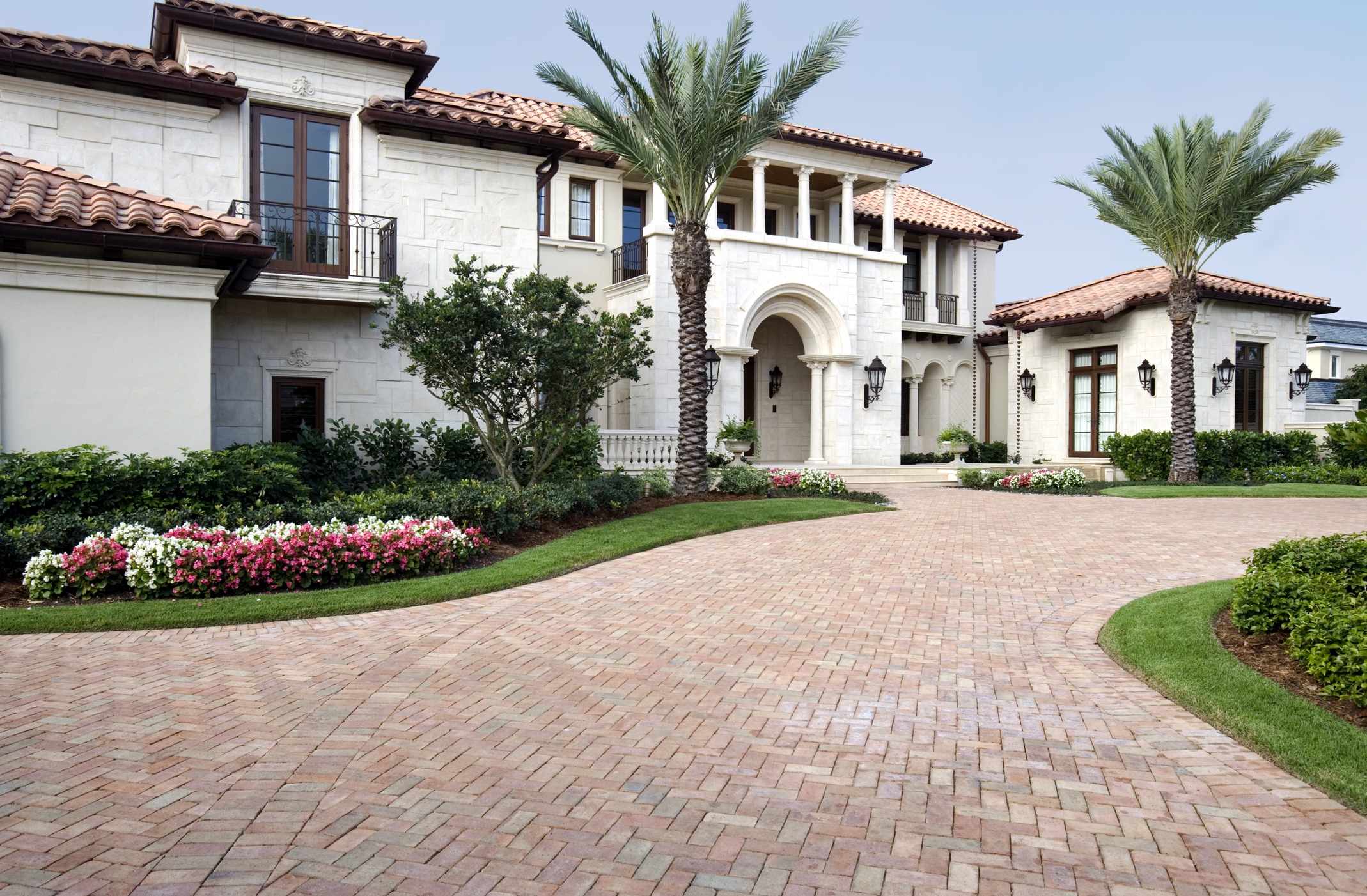 Mediterranean-style villa with stucco walls, arched entrance, red-tiled roof, manicured garden, and palm trees lining the brick-paved driveway.