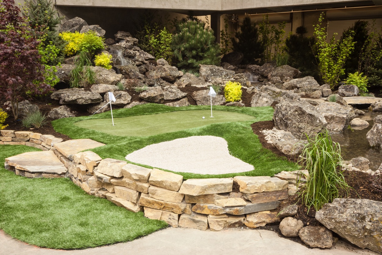 A small landscaped mini-golf course with a putting green, sand trap, and lush foliage surrounded by rocks and a retaining wall.