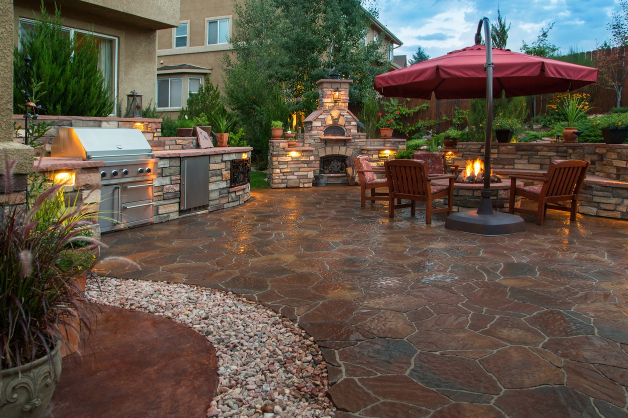 A cozy outdoor patio features a grill, fireplace, seating around a fire pit, large red umbrella, and lush greenery in a serene setting.