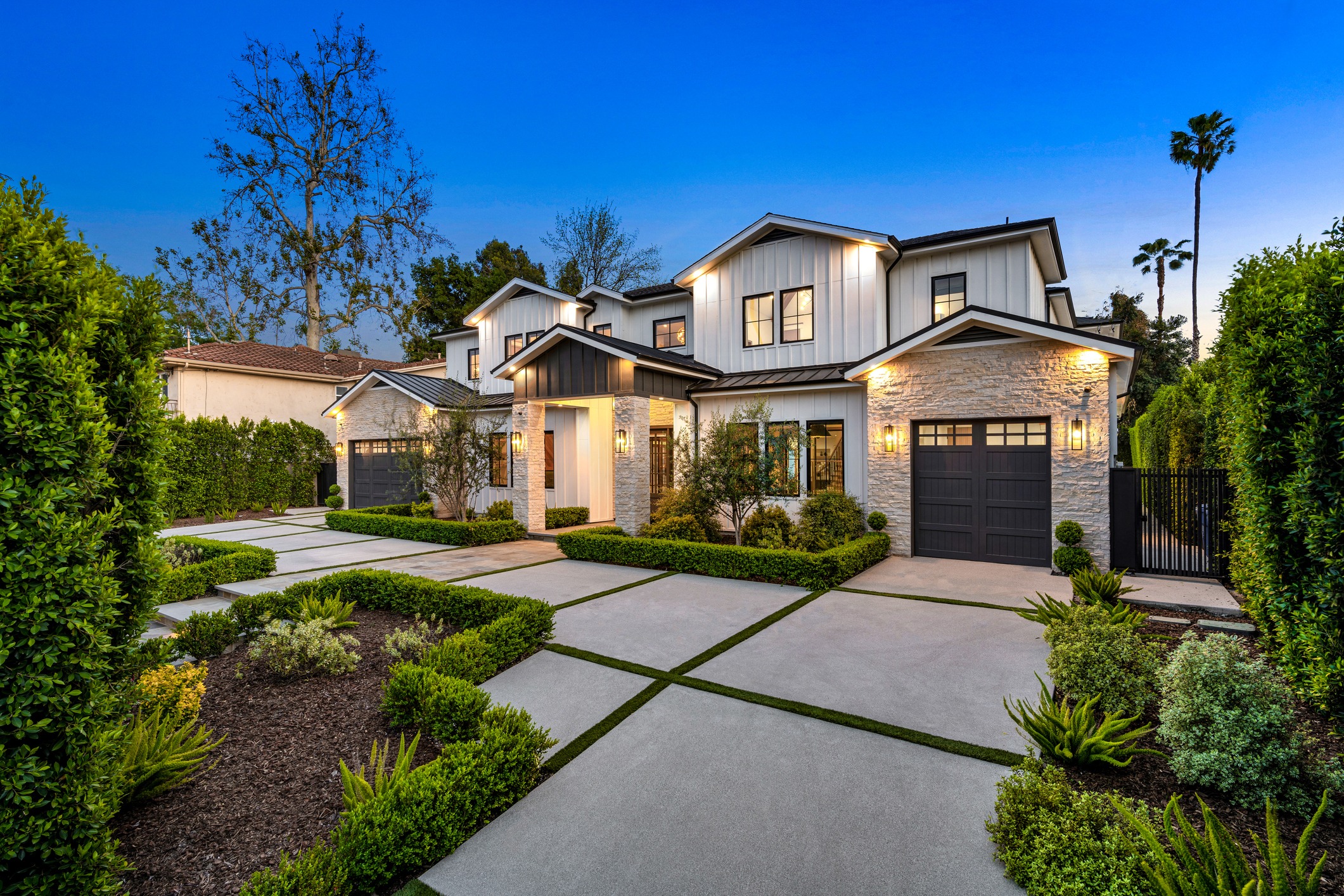 Modern house with lights on, surrounded by lush greenery and neatly trimmed hedges, during twilight. Spacious driveway and welcoming entrance pathway.