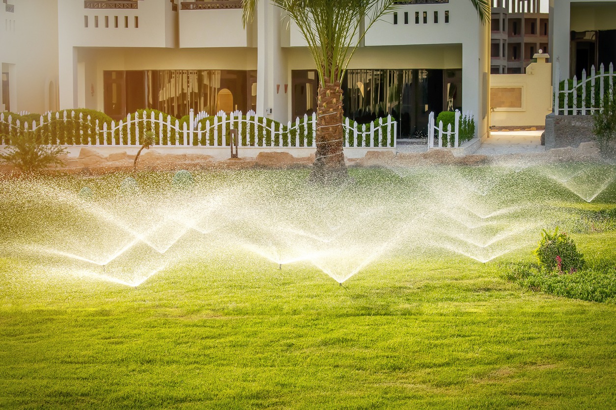 A lush garden with sprinklers showers green grass; white fencing and modern building in background enhance the serene residential setting.