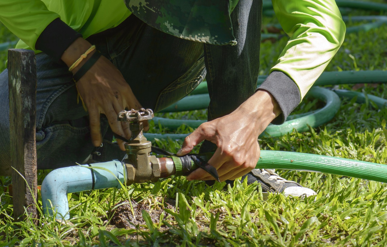A person in green clothing is fixing a hose on a grassy lawn, adjusting a blue pipe connection with focus and precision.