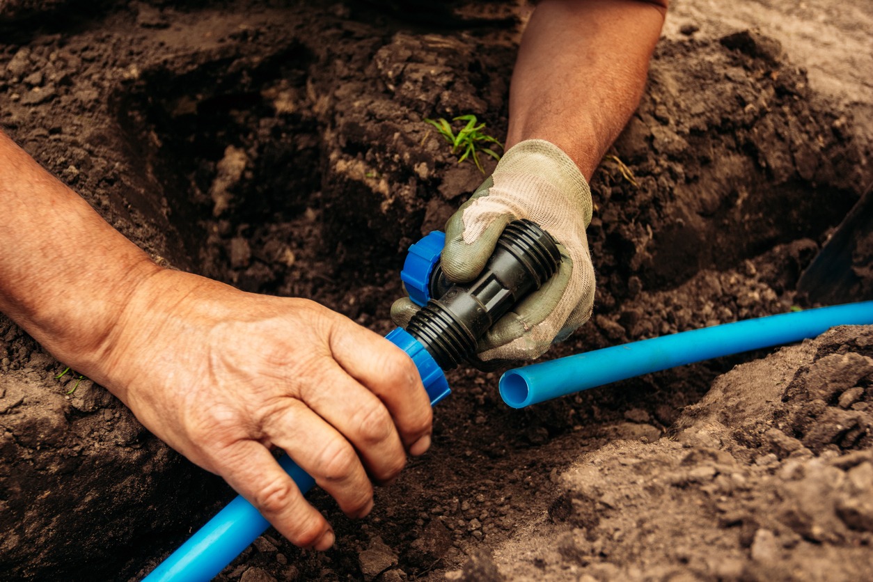 A person connects blue plastic pipes with a black fitting in a dirt trench, wearing gloves for protection and stability.