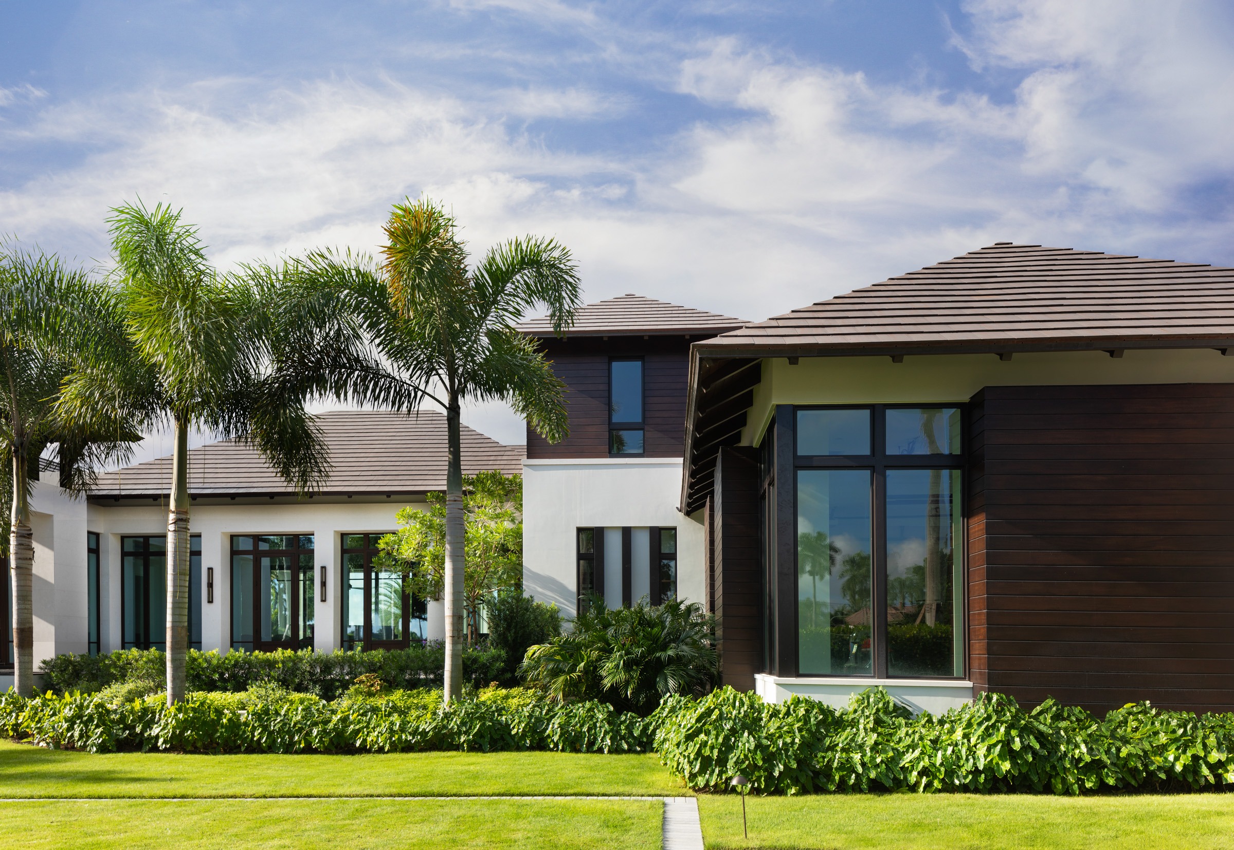 Modern house with large windows, surrounded by lush greenery and towering palm trees against a blue sky, creating a tropical, serene atmosphere.
